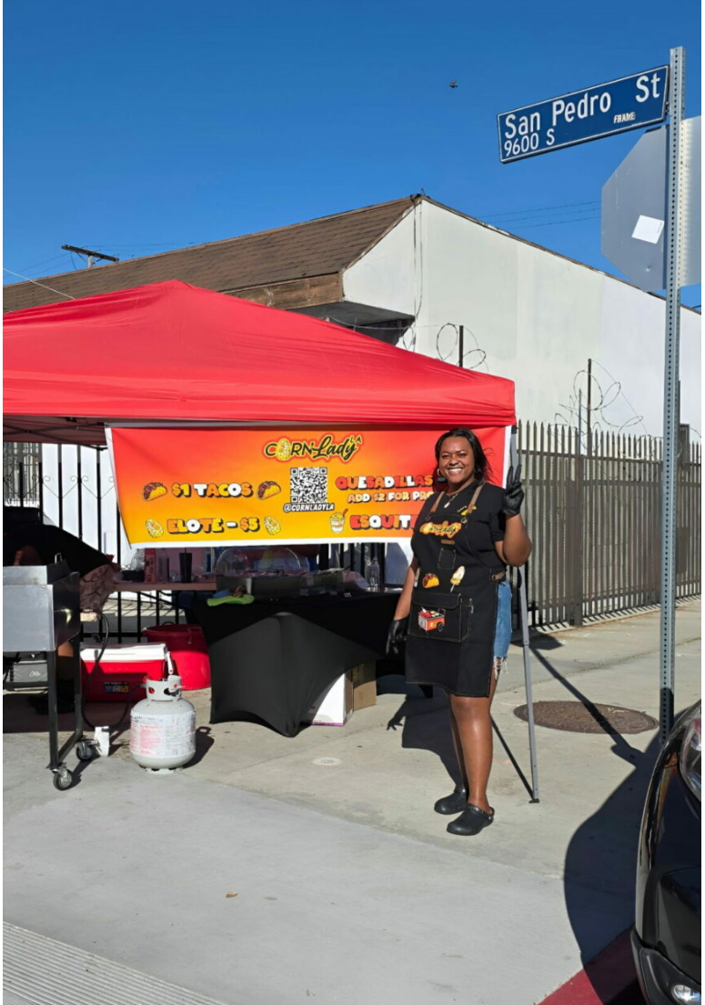 The owner of Corn Lady LA standing at her red canopy stand on San Pedro Street